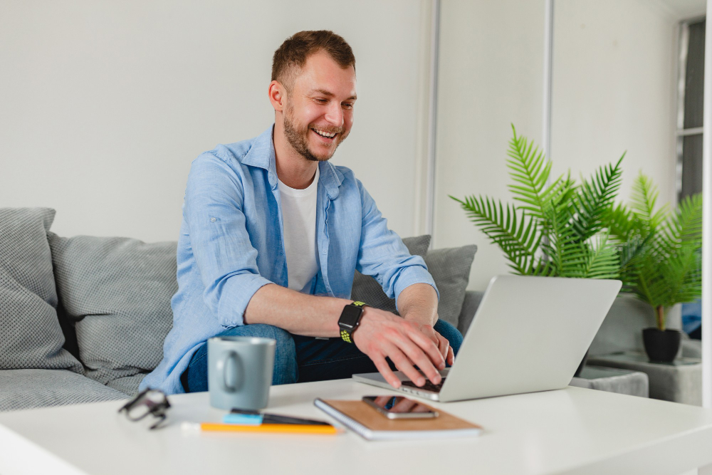 handsome-smiling-man-sitting-sofa-drinking-tea-home-table-working-online-laptop-from-home